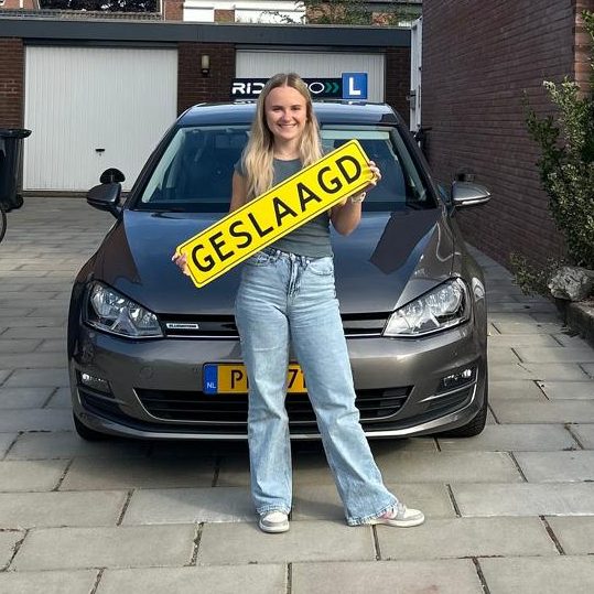 Blonde vrouw houdt een verkeersbord met de tekst "GESLAAGD" voor een auto.